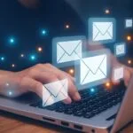 Nahaufnahme einer Person, die auf einem Laptop tippt, während leuchtende E-Mail-Symbole über der Tastatur schweben. Umgeben von blauen und orangefarbenen Lichtpunkten, die eine digitale Kommunikation symbolisieren.